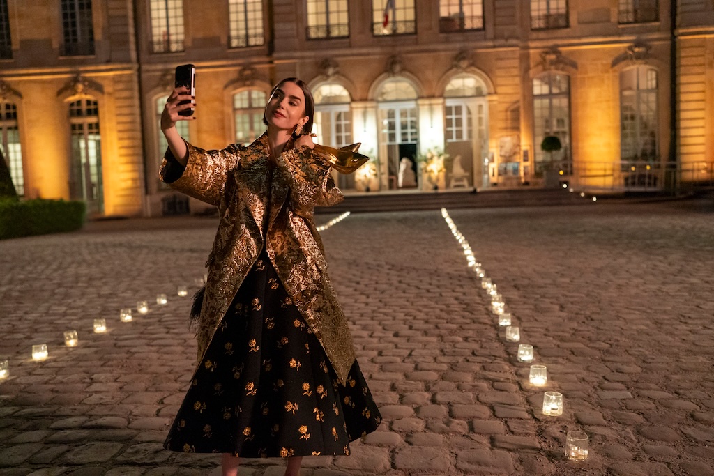 "Woman in a gold coat and black dress takes a selfie with her phone at night in a candlelit courtyard outside an elegant, well-lit building with large windows and cobblestone ground."